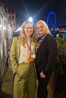 Elizabeth Forsythe in green trouser suit and Kerry Leeson Beevers in black trousers suit both with blonde hair on the terrace at the Houses of Parliament with the London Eye in the background