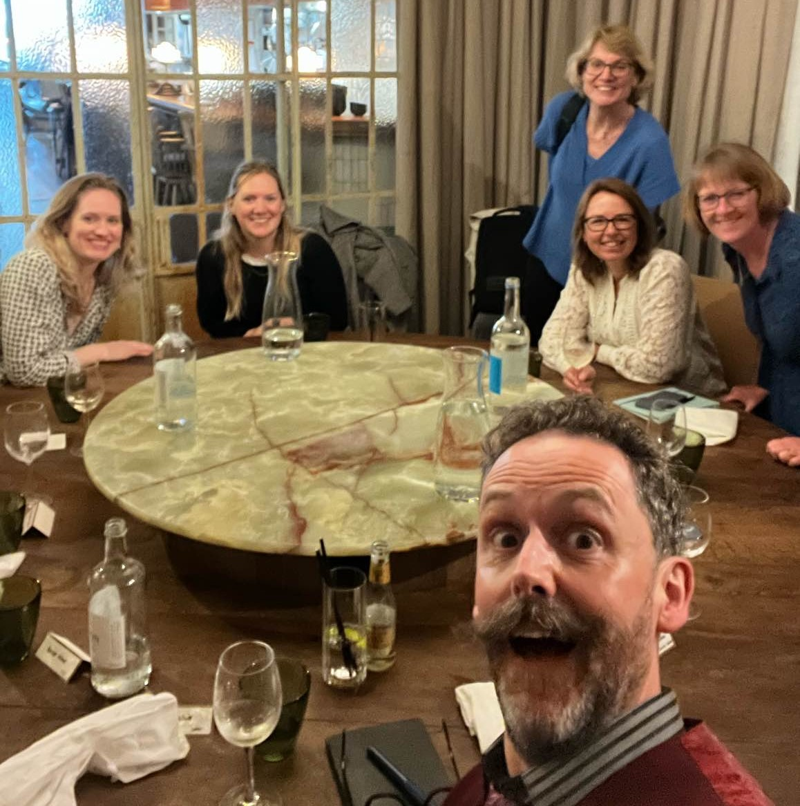 Five women and one man (taking the selfie) around a round table all smiling.